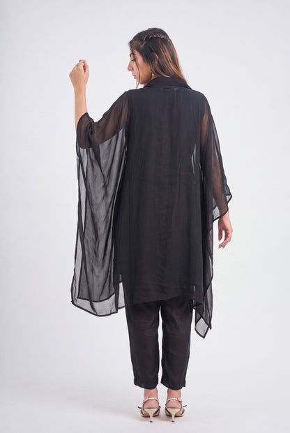 A woman shown from the back wearing a sheer black long tunic or kaftan with wide draped sleeves, paired with black pants and beige heeled sandals, against a plain light gray studio background.
