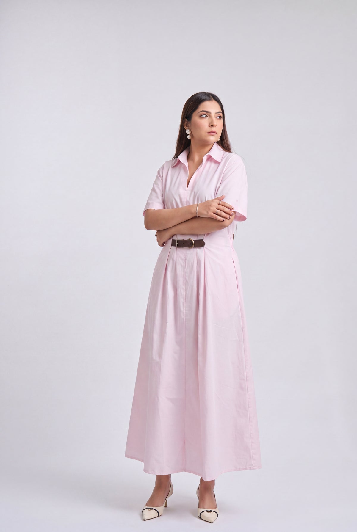A woman modeling a light pink short-sleeve midi shirt dress with a black belt, holding a beige tote bag and wearing beige heels, against a light gray studio background.