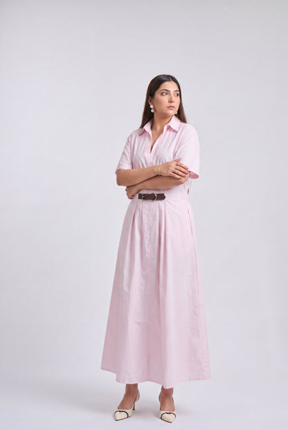 A woman modeling a light pink short-sleeve midi shirt dress with a black belt, holding a beige tote bag and wearing beige heels, against a light gray studio background.
