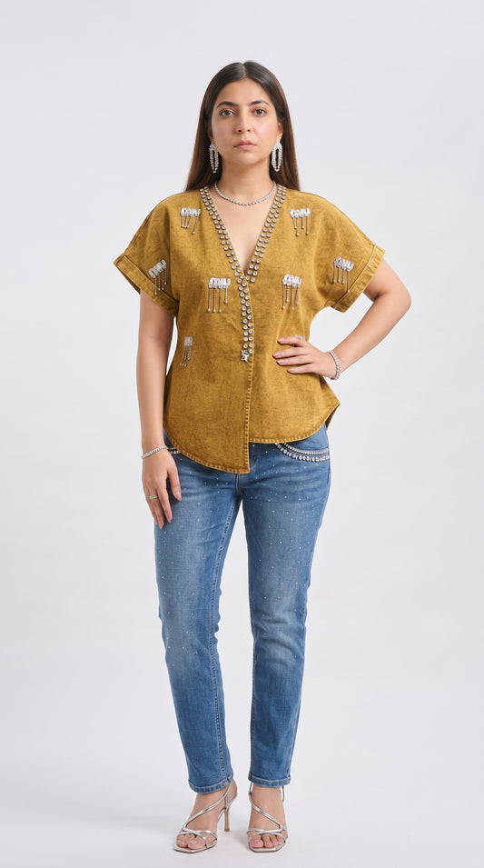 Woman wearing a mustard yellow top with embellishments and blue jeans on a white background