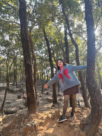 Person standing in a forest with arms outstretched, wearing a light blue jacket with a red bow.