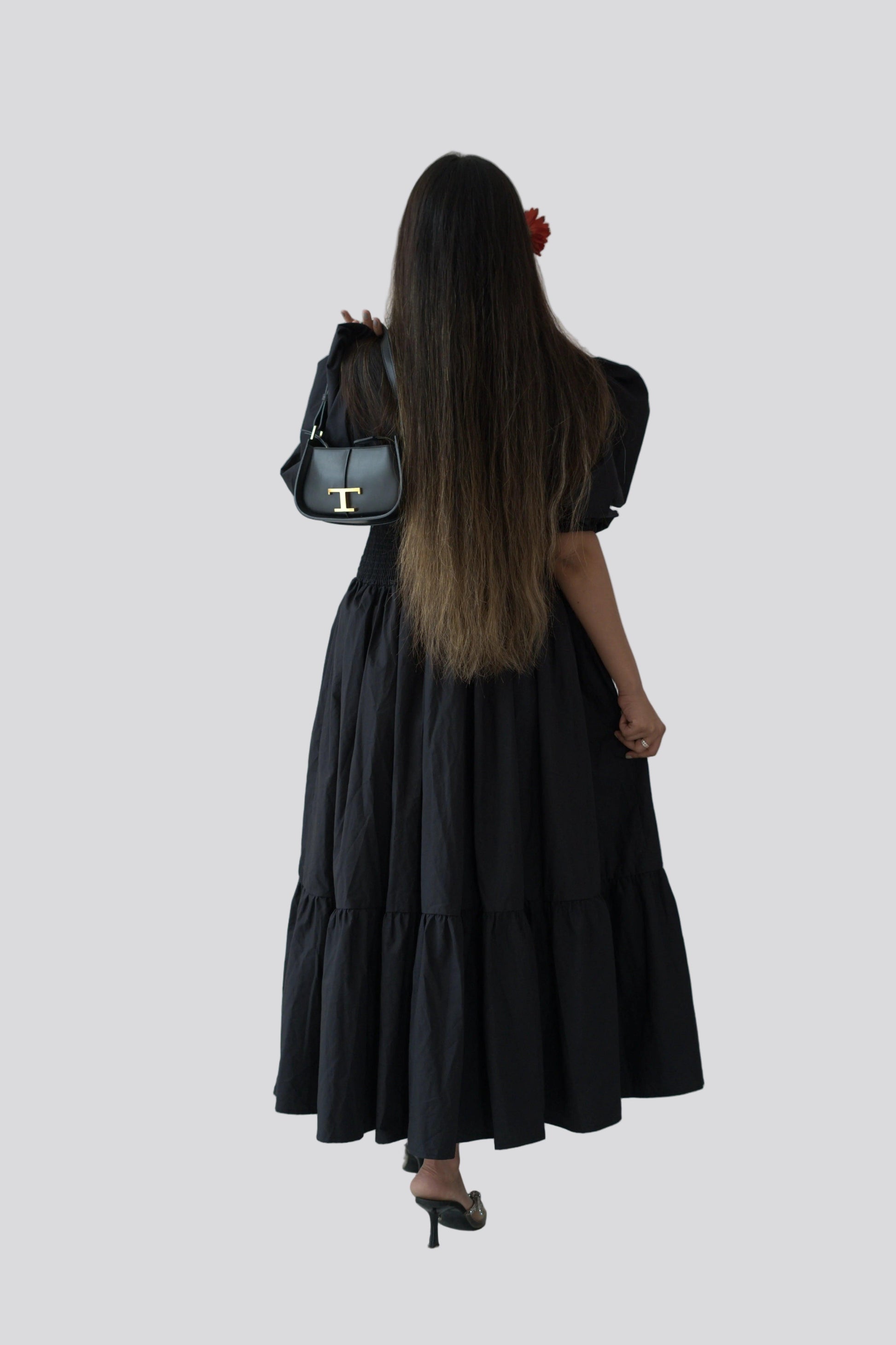 Person wearing a black dress holding a black handbag against a white wall.
