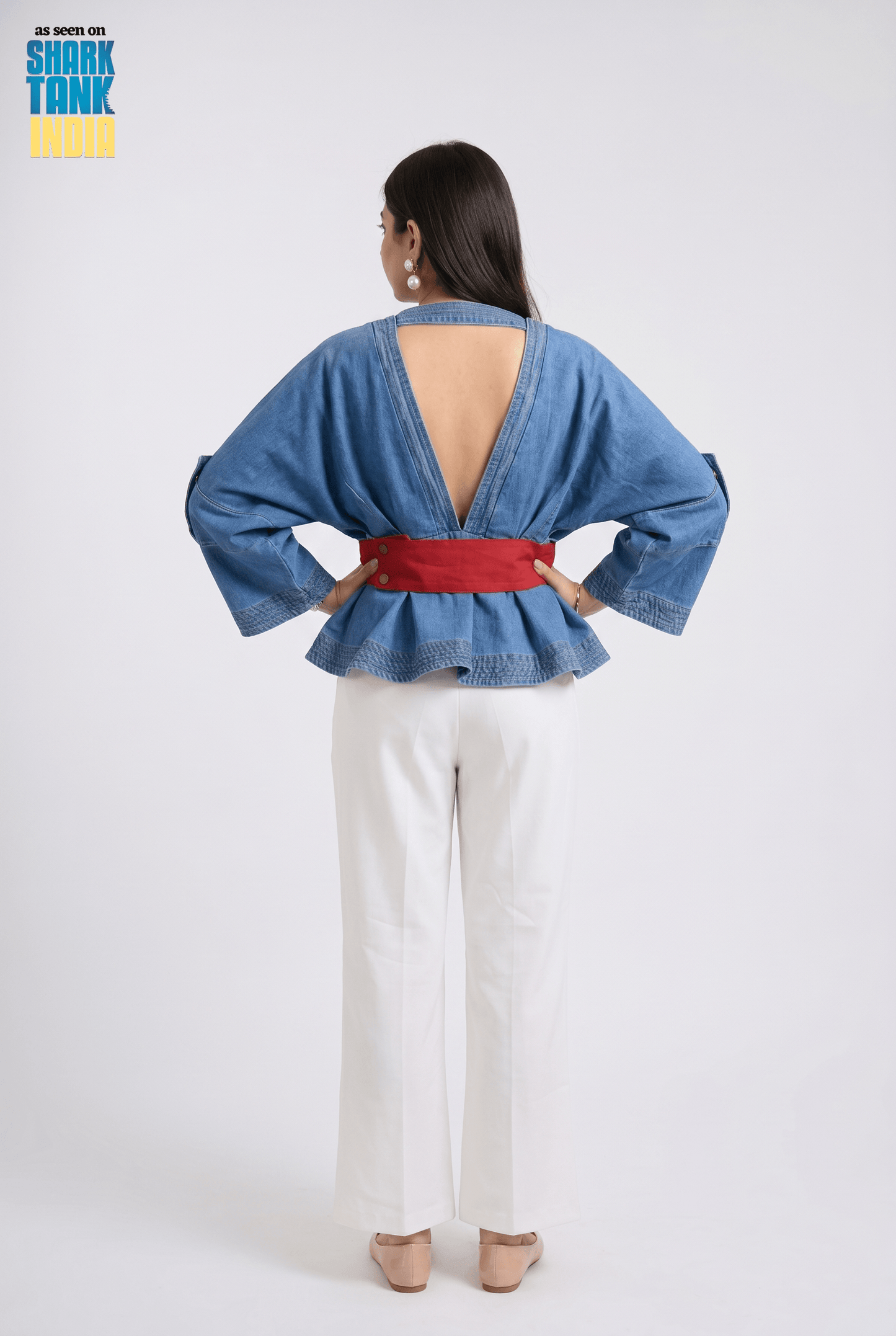 Woman wearing a blue denim jacket with a red belt and white pants on a plain background.