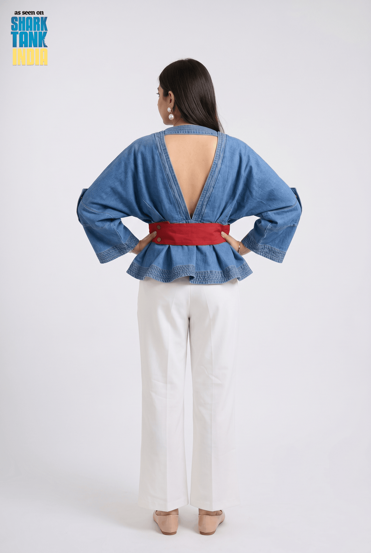 Woman wearing a blue denim jacket with a red belt and white pants on a plain background.