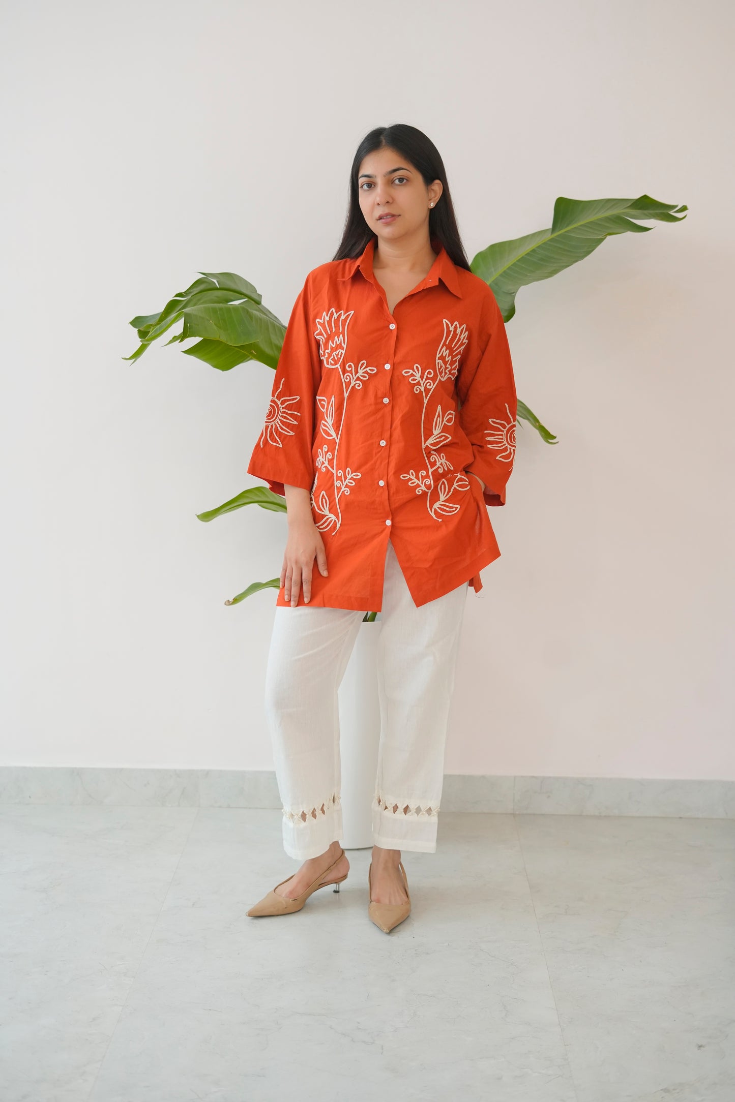 Woman wearing an orange embroidered shirt and white pants standing against a white wall with a plant.