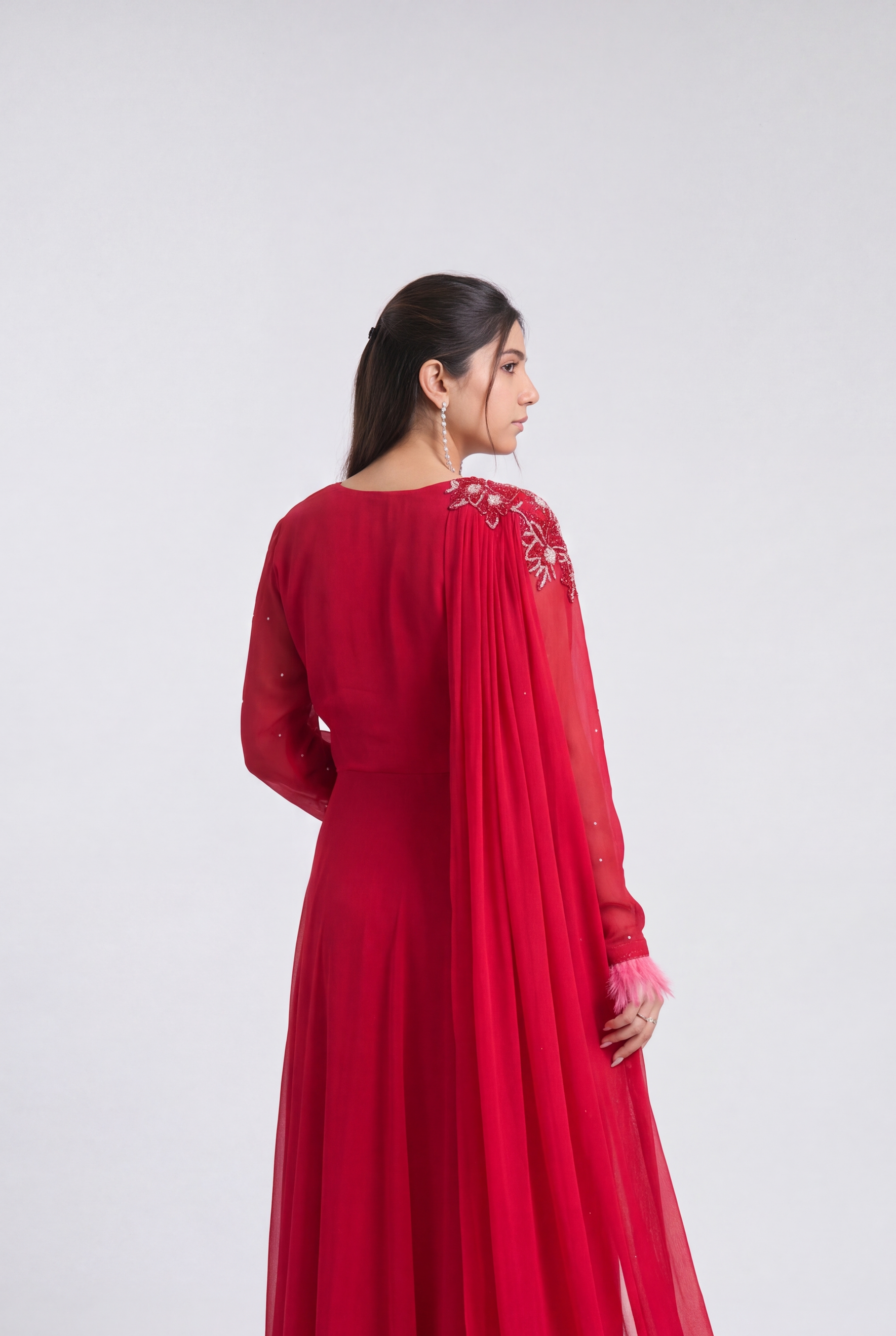 A woman wearing a long-sleeved red gown with a sheer draped overlay and feathered hem, photographed from the back against a plain light gray studio background.