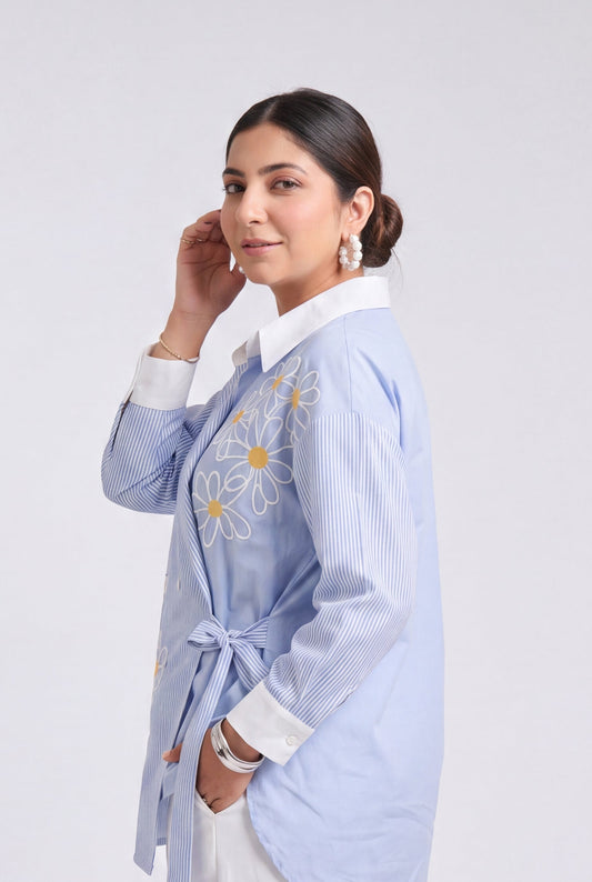 A woman modeling a light blue long-sleeve button-up shirt with white collar and cuffs, featuring yellow and orange floral embroidery on the chest, paired with white trousers and clear high-heeled sandals against a light gray studio background.