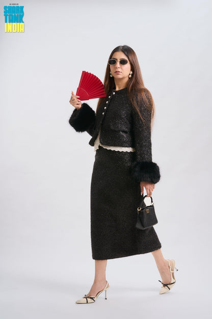 Woman in a stylish outfit with a black coat, skirt, and fur trim holding red fans against a white background.