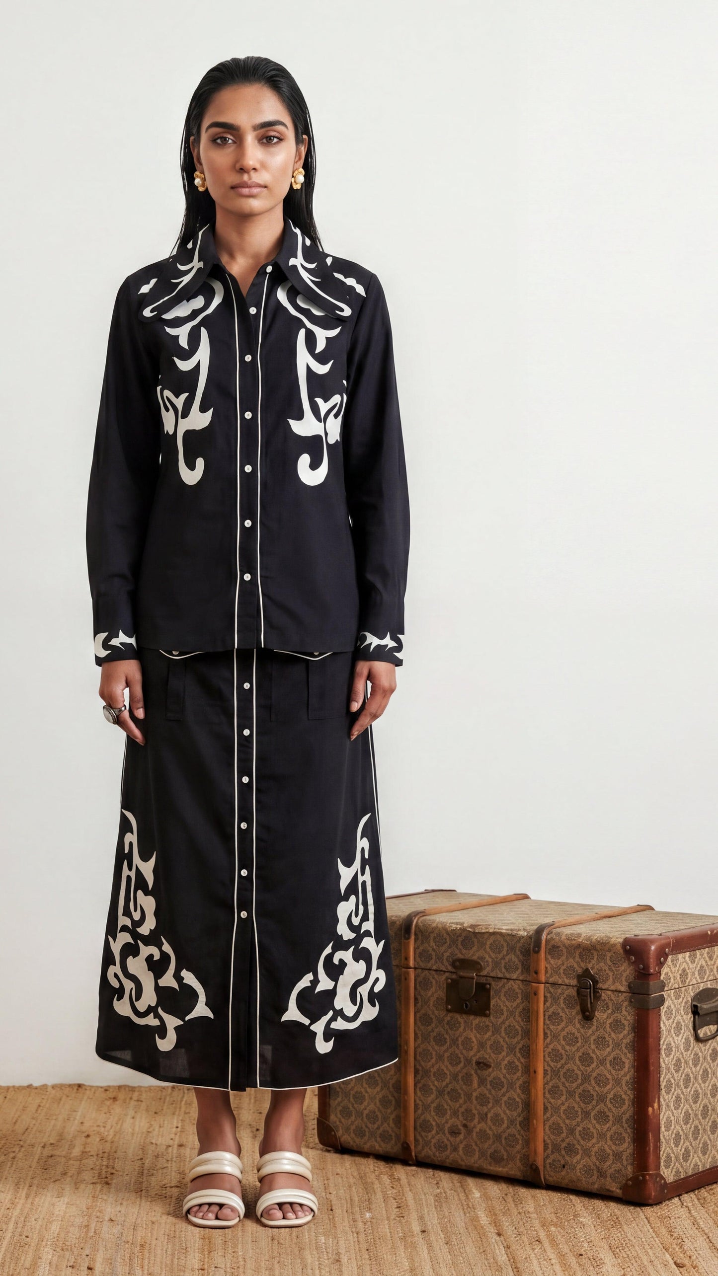 Woman wearing a black outfit with white embroidery standing next to a vintage suitcase.
