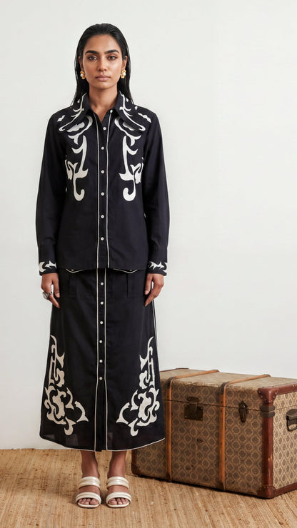 Woman wearing a black outfit with white embroidery standing next to a vintage suitcase.