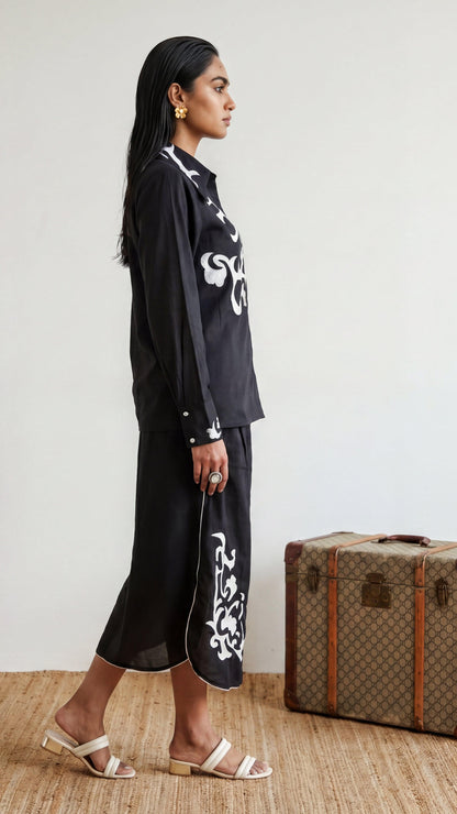 Woman in black outfit with white patterns standing next to a vintage suitcase.