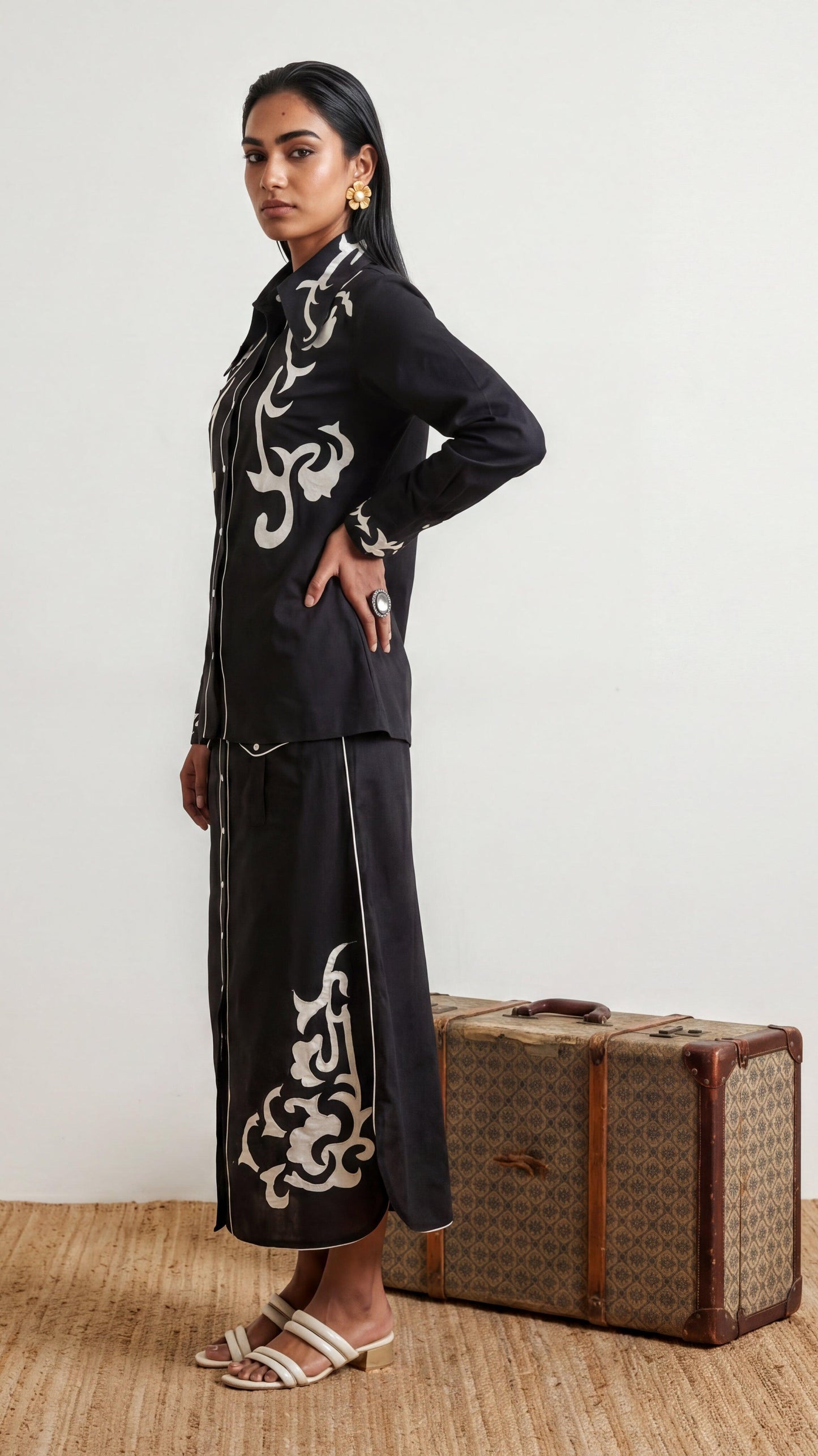 Woman in black outfit with white patterns standing next to a vintage suitcase on a wooden floor.