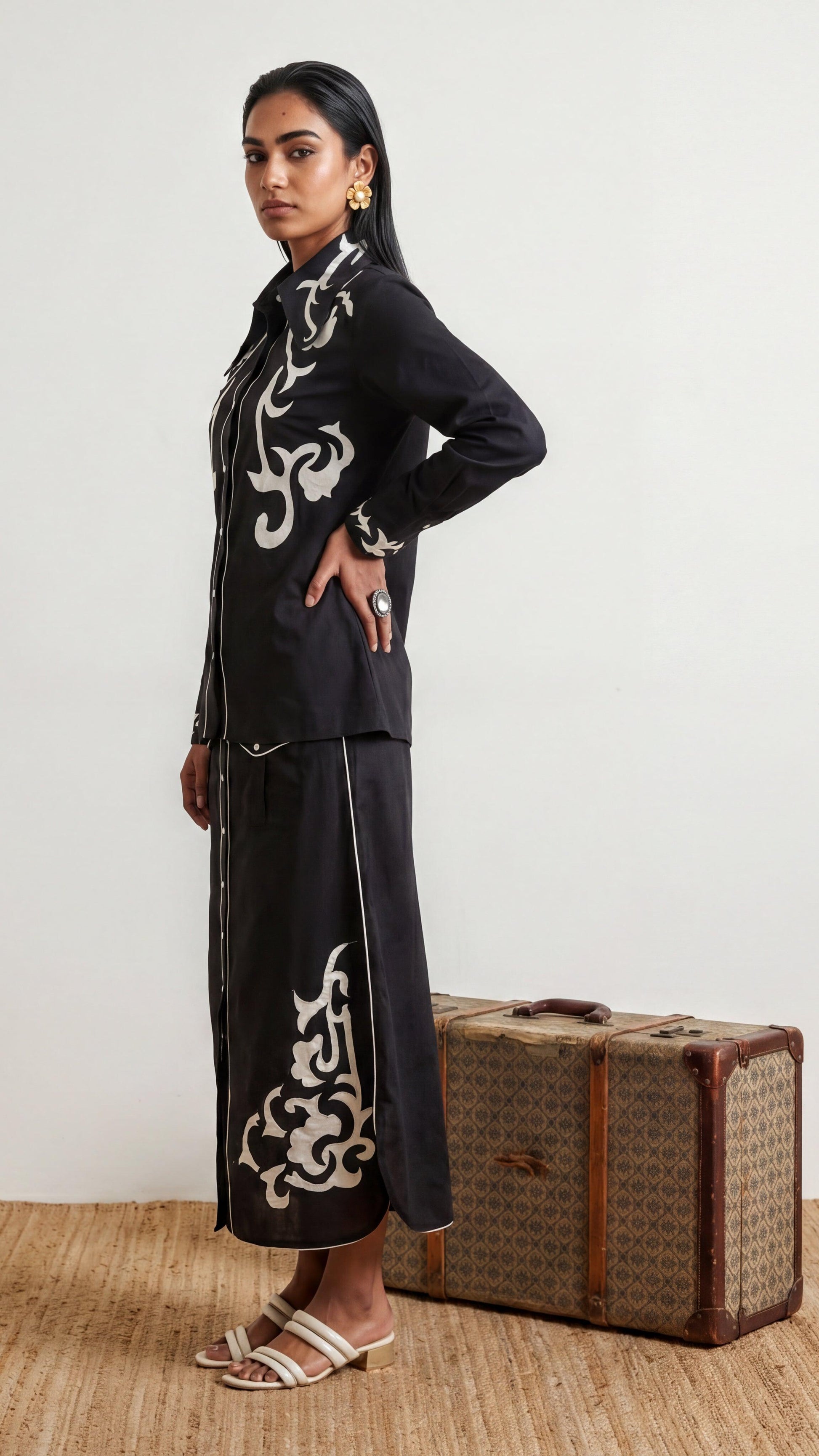 Woman in black outfit with white patterns standing next to a vintage suitcase on a wooden floor.