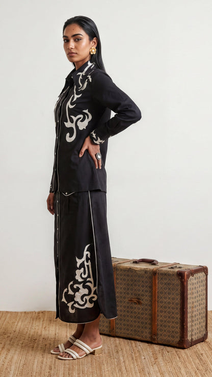 Woman in black outfit with white patterns standing next to a vintage suitcase on a wooden floor.