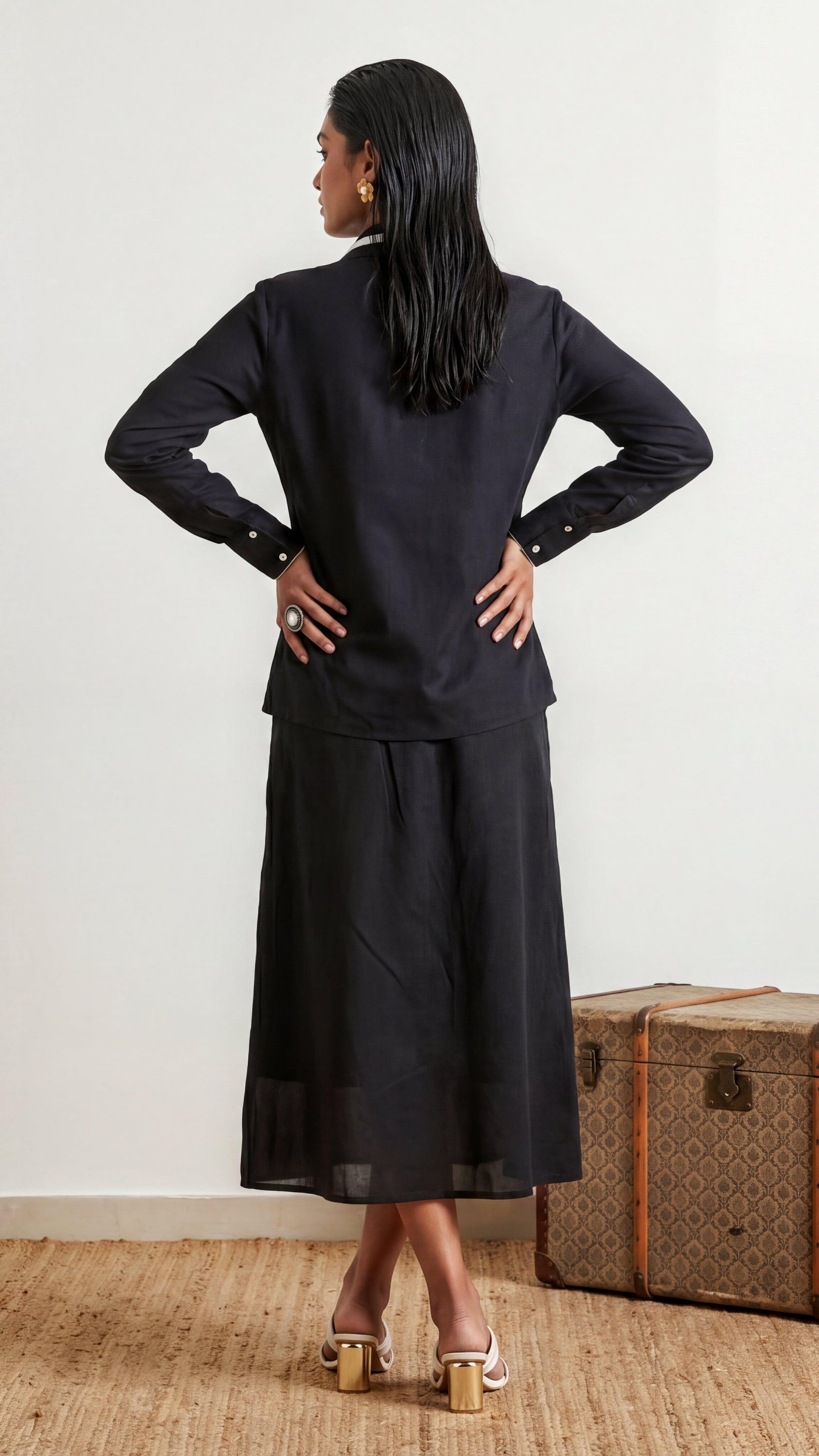 Woman wearing a black outfit standing next to a vintage suitcase on a wooden floor.