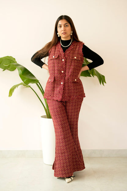 Woman wearing a red checkered outfit with a vest and pants, standing against a white wall with a plant.