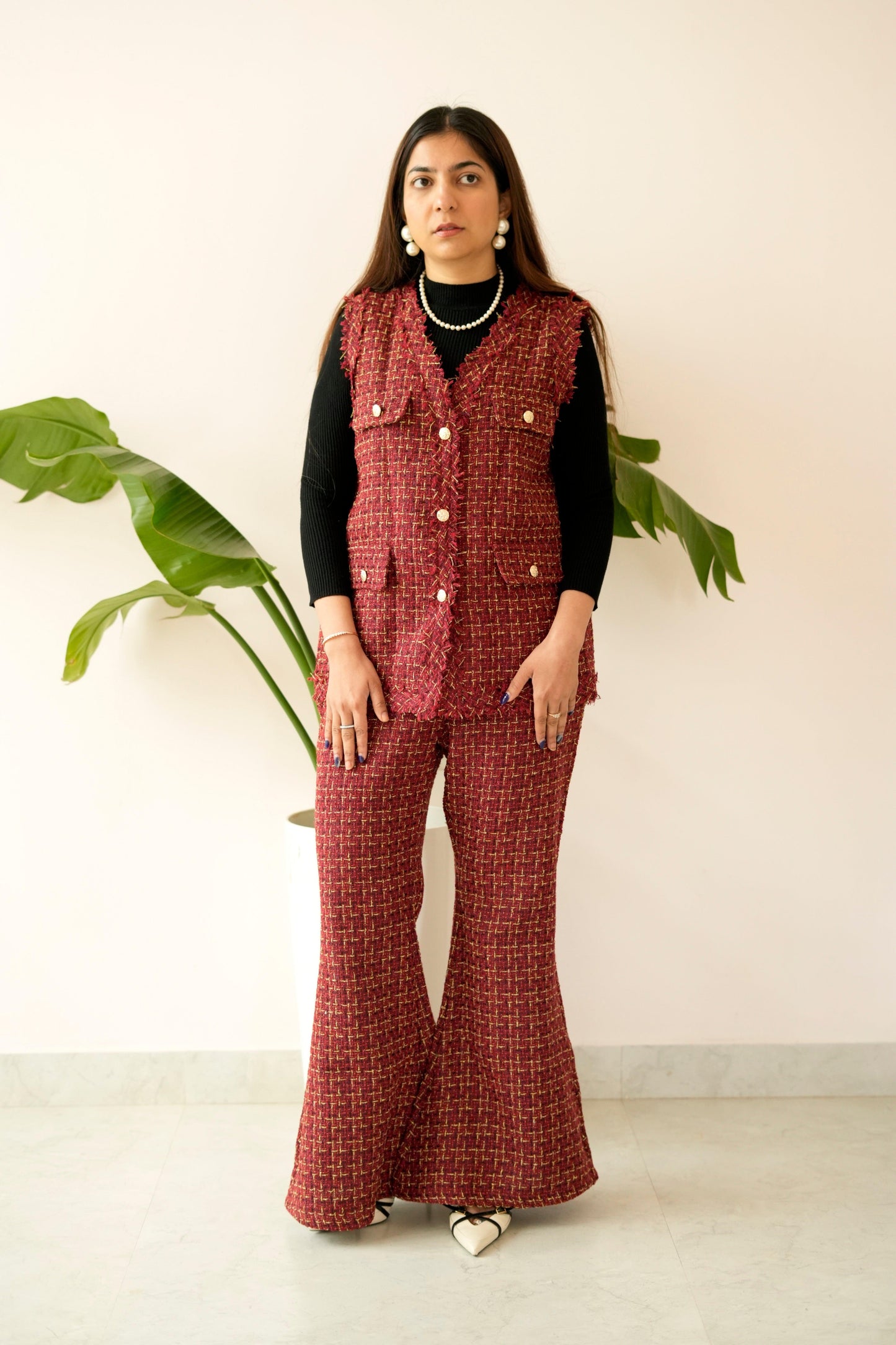 Woman wearing a red checkered vest and flared pants standing against a white wall with a plant.