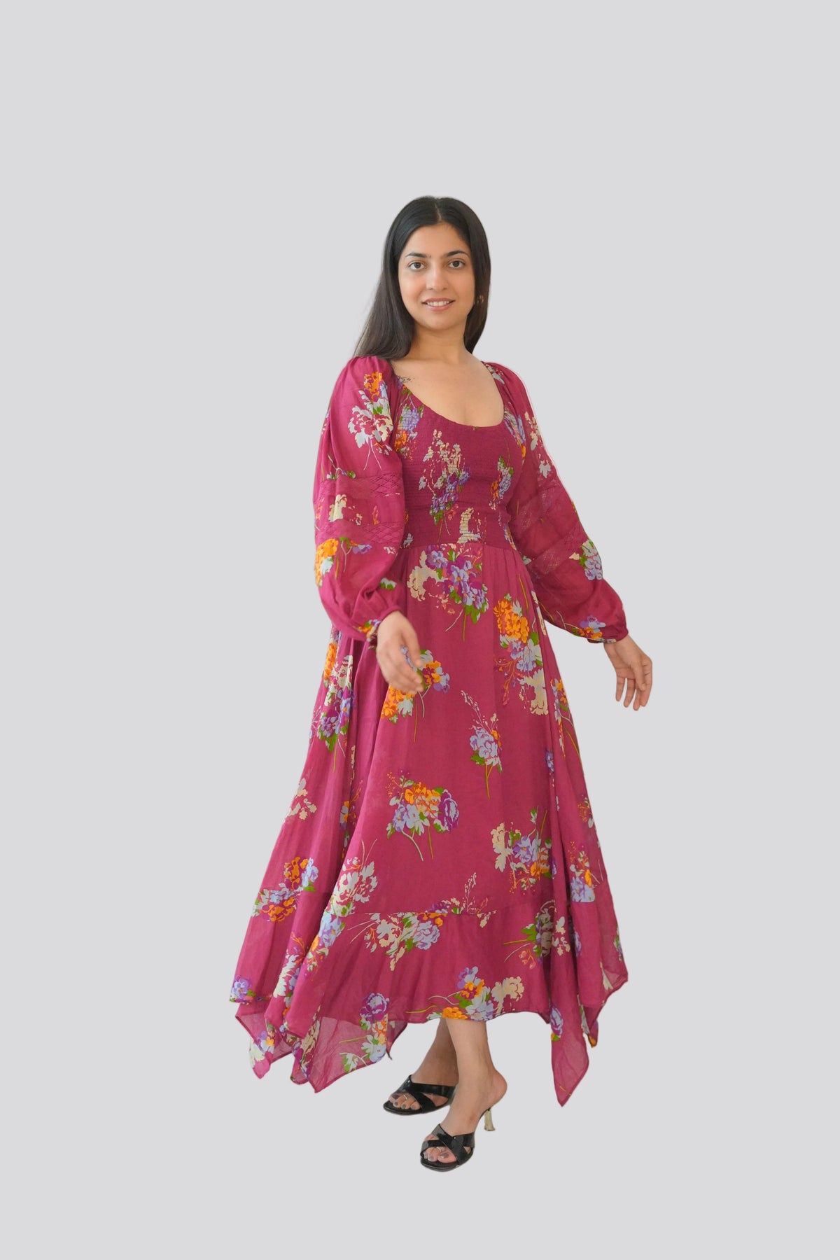 Woman in a pink floral dress standing next to a large green plant against a white wall.