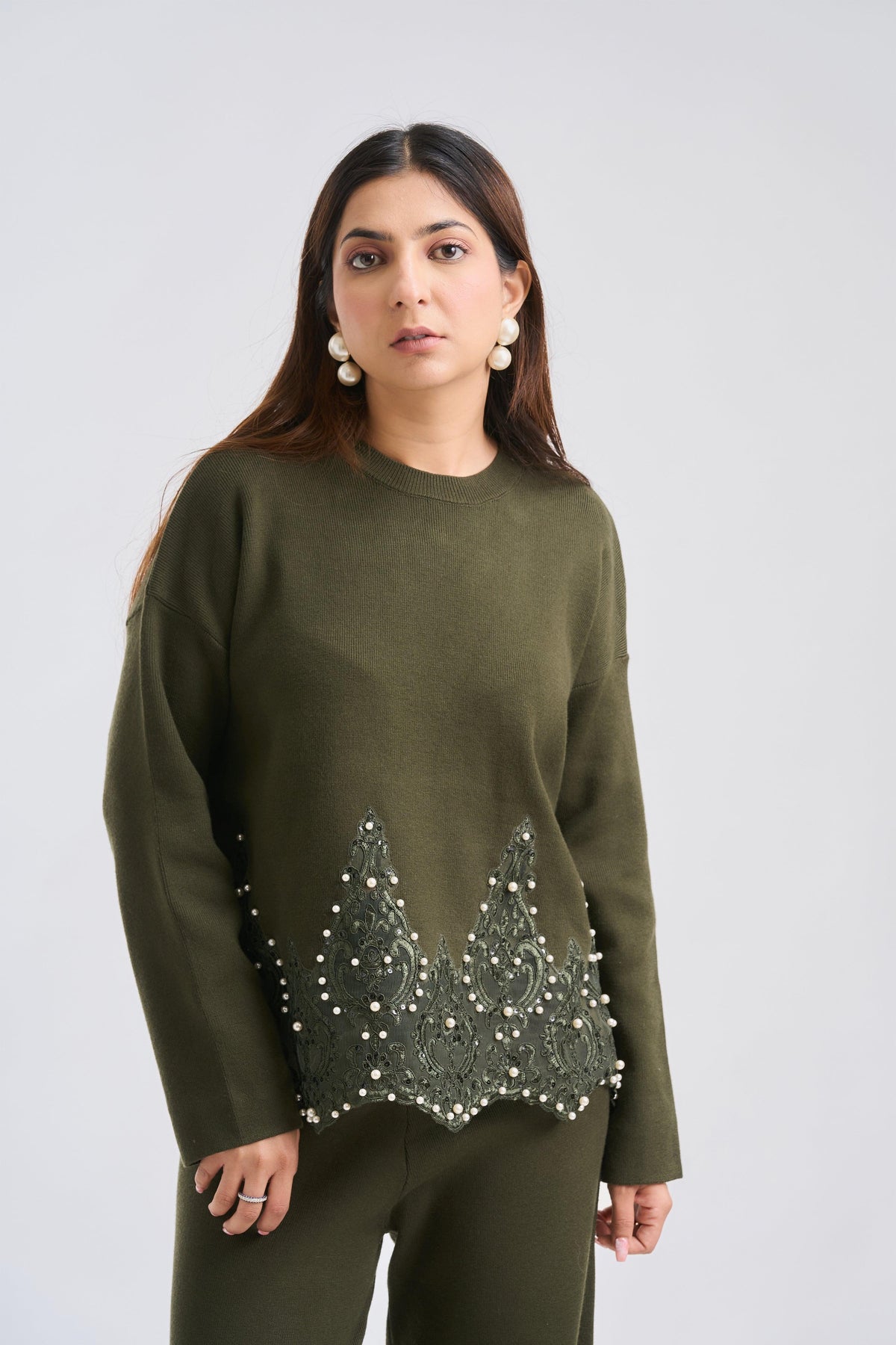 Woman wearing an olive green sweater with bead embellishments on a plain background
