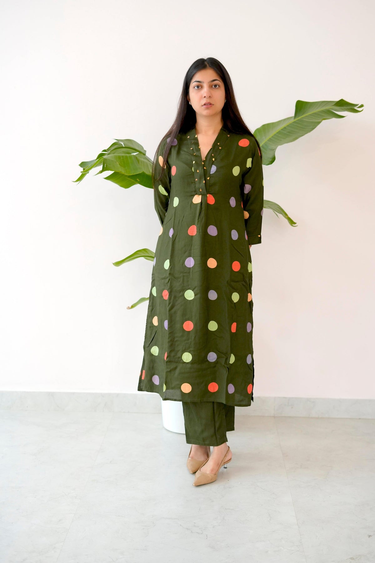 Woman wearing a green outfit with colorful polka dots in front of a white wall with a plant.