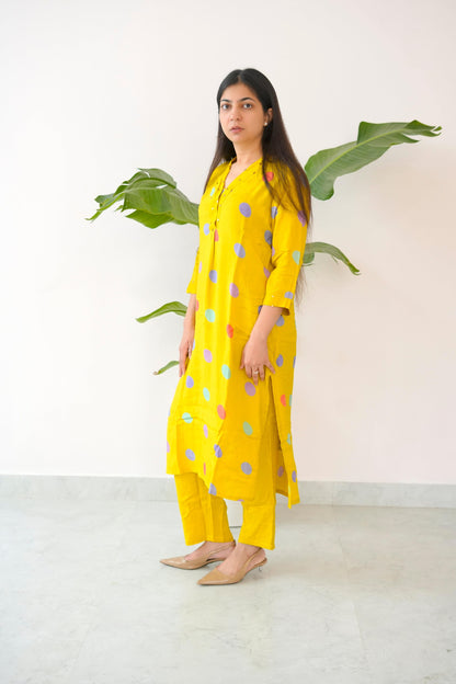 Woman wearing a yellow outfit with colorful polka dots standing against a white wall with green leaves.