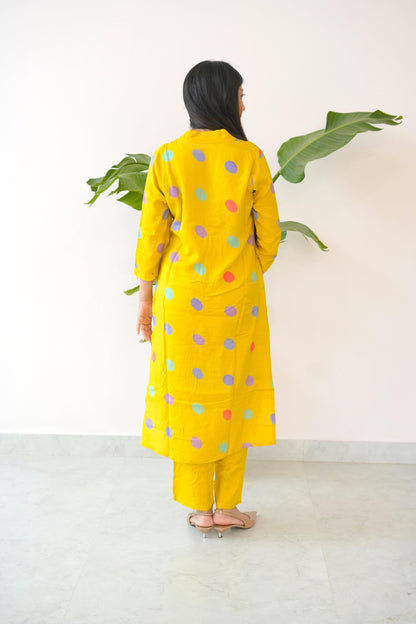 Person wearing a yellow outfit with colorful polka dots, standing against a white wall with green leaves.