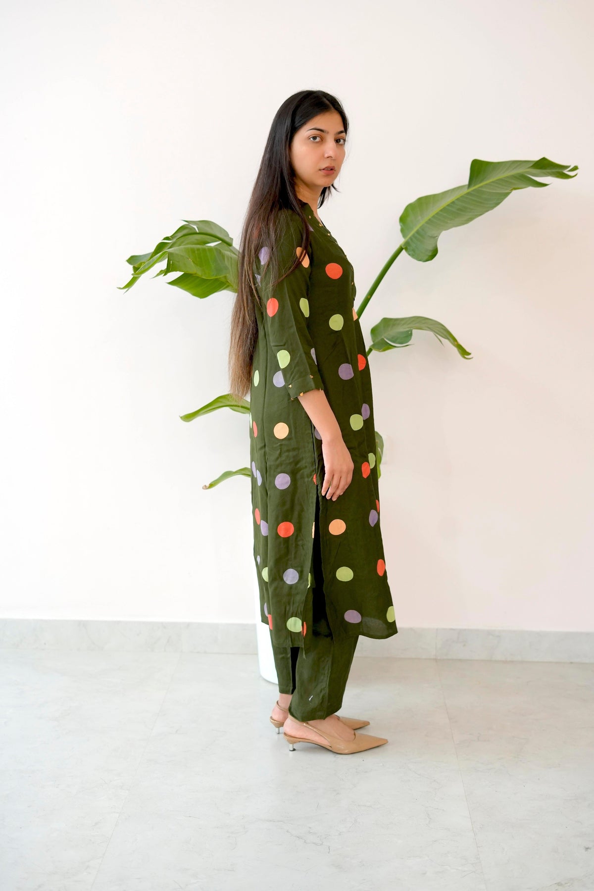 Woman wearing a green polka dot dress standing next to a plant indoors.