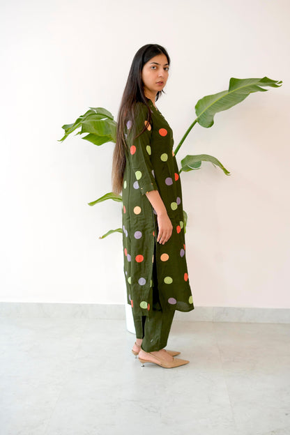 Woman wearing a green polka dot dress standing next to a plant indoors.