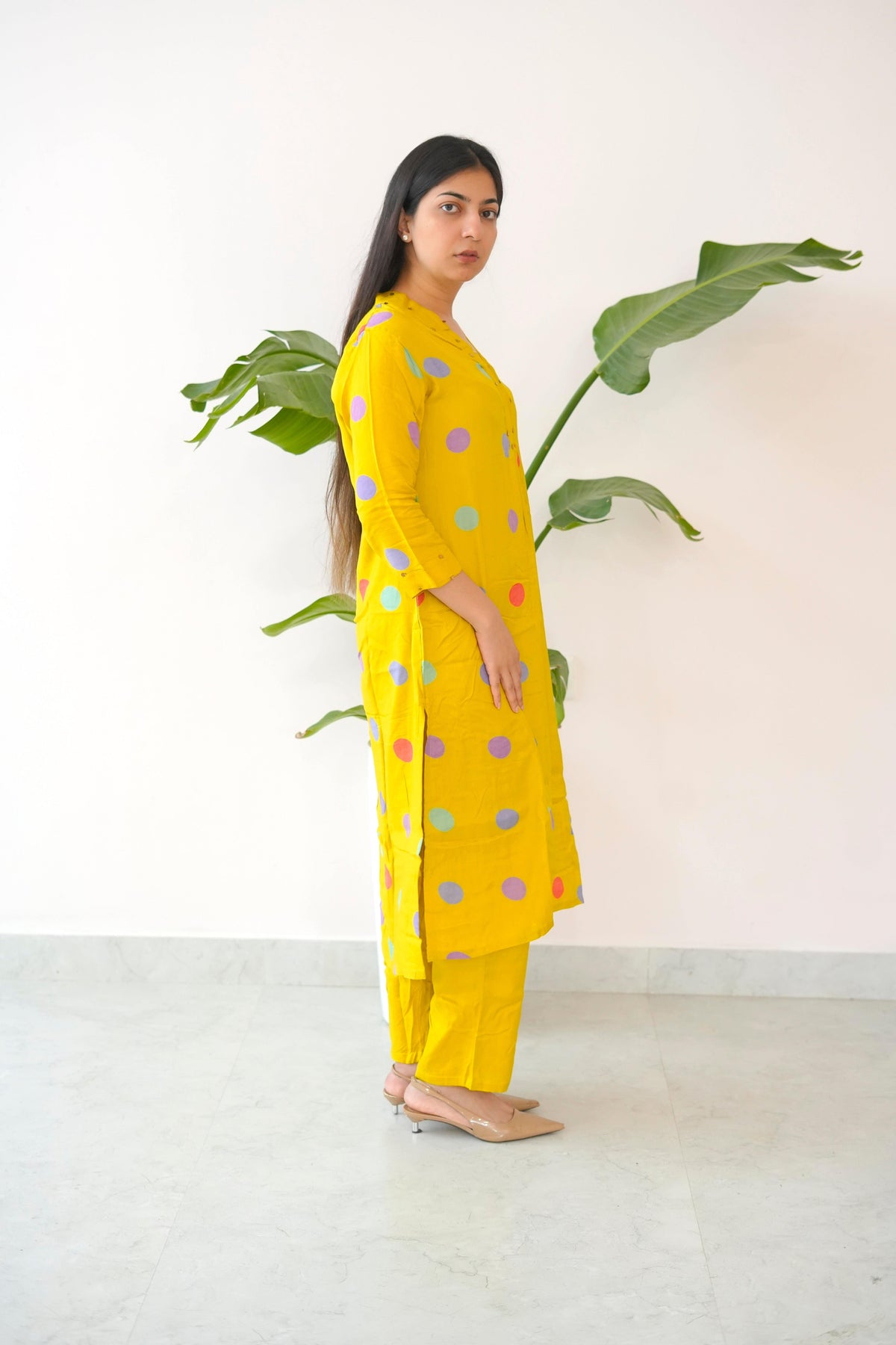 Woman wearing a yellow outfit with colorful polka dots standing next to a plant indoors.