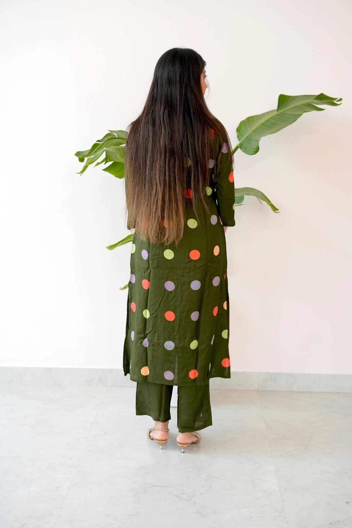 Woman wearing a green outfit with colorful polka dots, standing in front of a white wall with a plant.