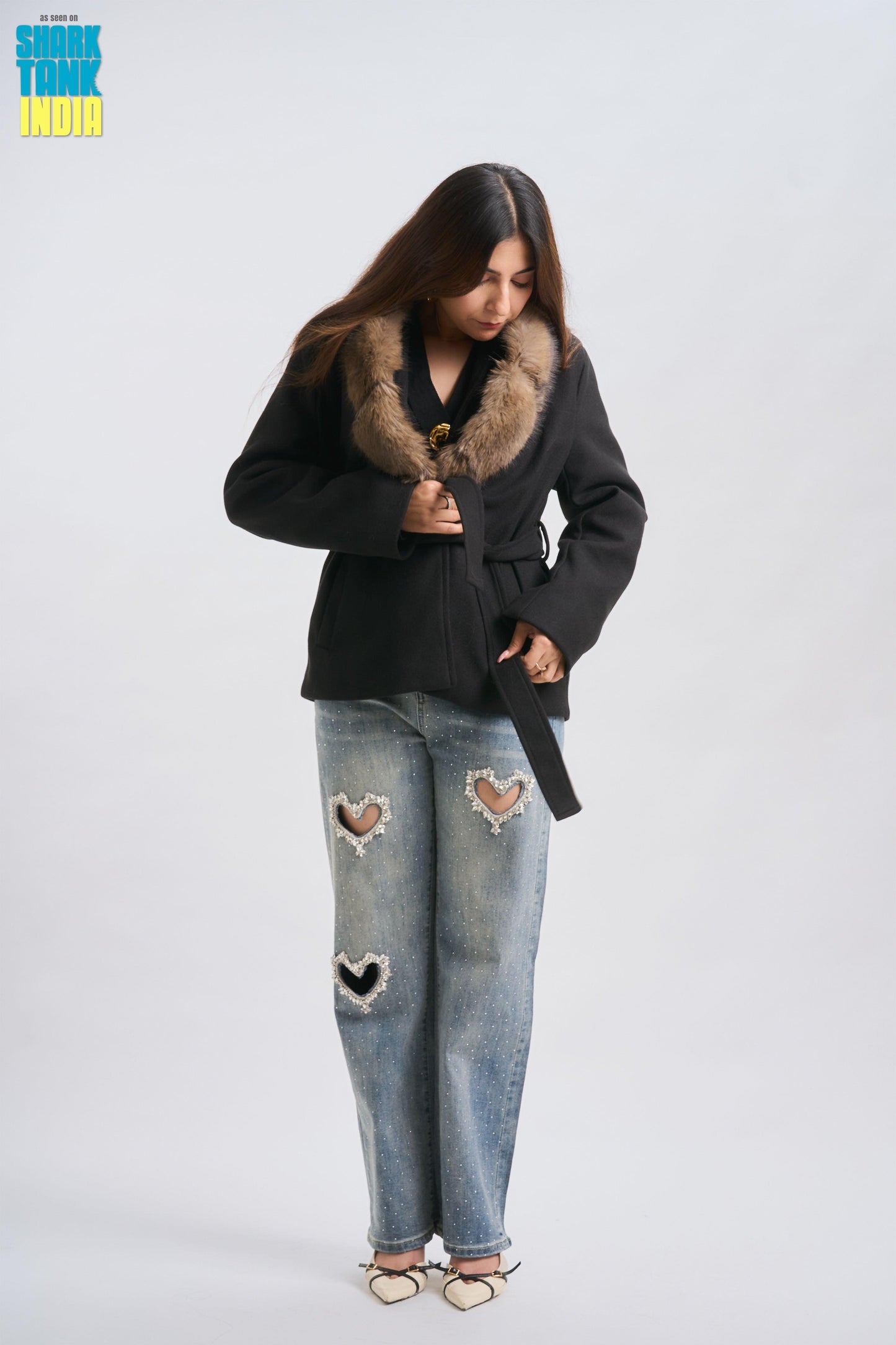 Person wearing a black coat with fur collar and ripped jeans on a white background