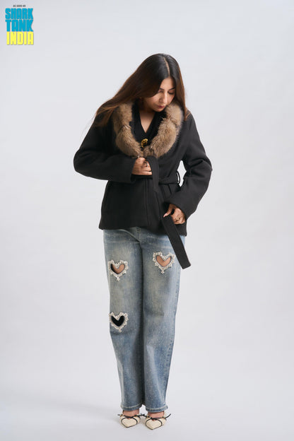Person wearing a black coat with fur collar and ripped jeans on a white background