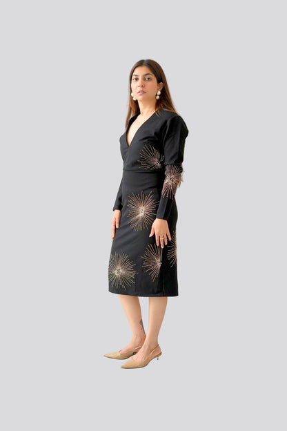 Woman wearing a black dress with gold patterns standing in front of a white wall with a plant.
