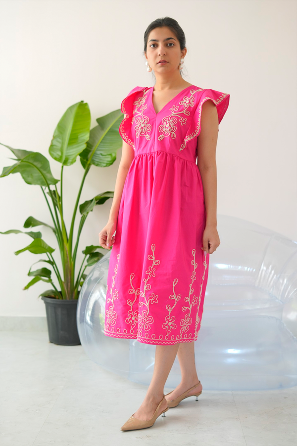 A woman standing in a room with a plant beside her, wearing a vibrant pink dress with intricate cream floral embroidery, flutter sleeves, and a V-neckline.