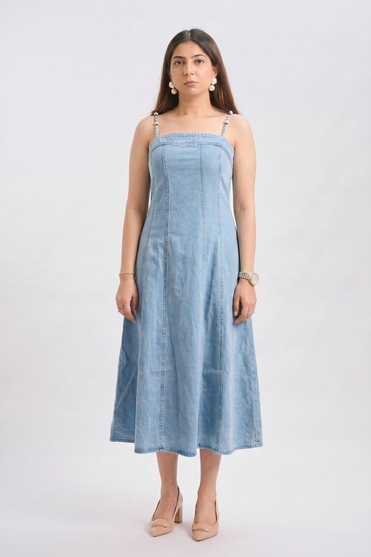 A woman modeling a light blue denim midi dress with thin shoulder straps, a smocked bodice, and an A-line skirt, standing against a plain light gray studio background.