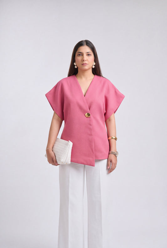 A woman modeling a pink short-sleeve wrap-style top with a single gold button, paired with white wide-leg pants and holding a white textured clutch, against a light gray studio background.