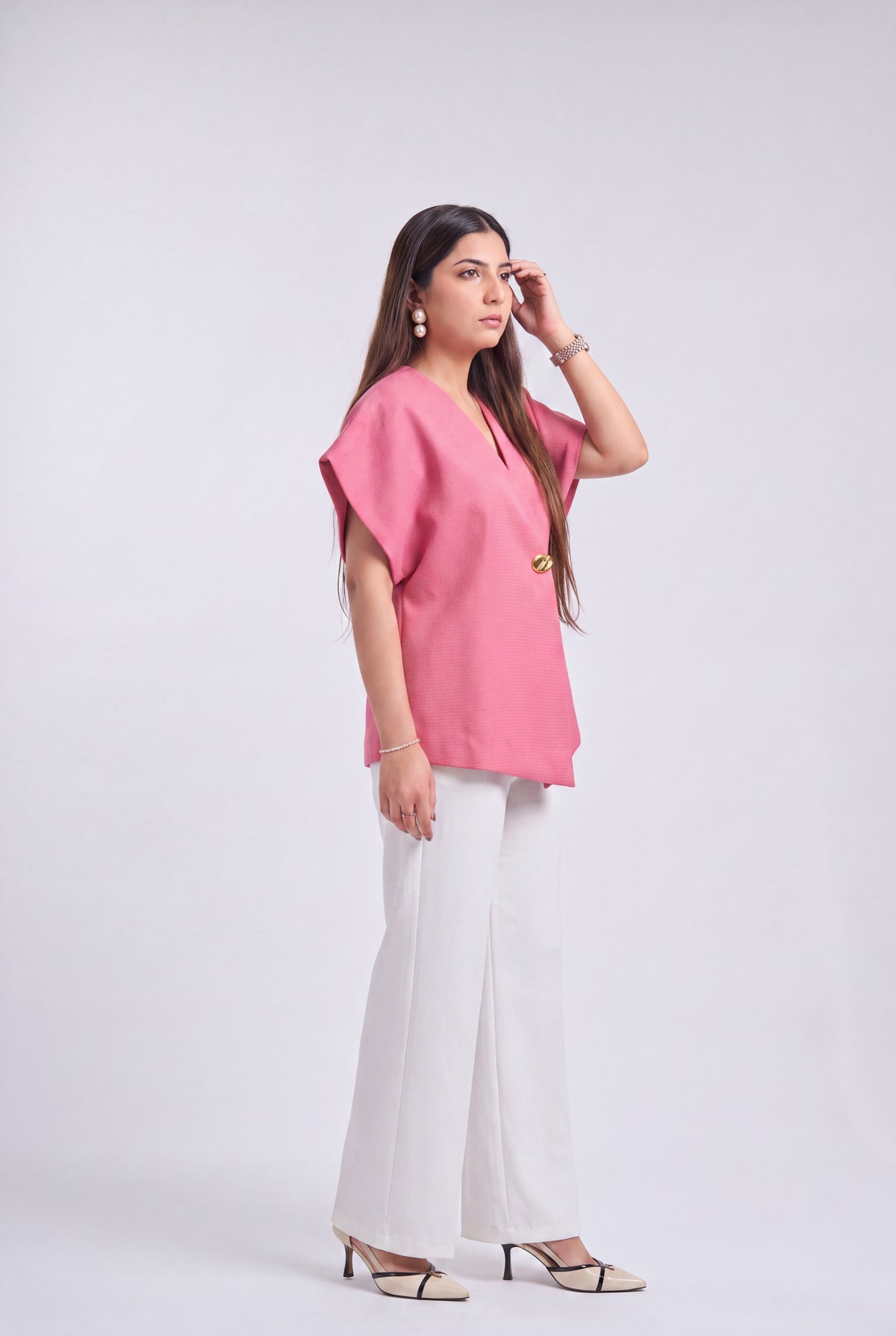 A woman modeling a pink short-sleeve top with a V-neck and gold button detail, paired with white wide-leg pants and beige heeled sandals, against a light gray studio background.