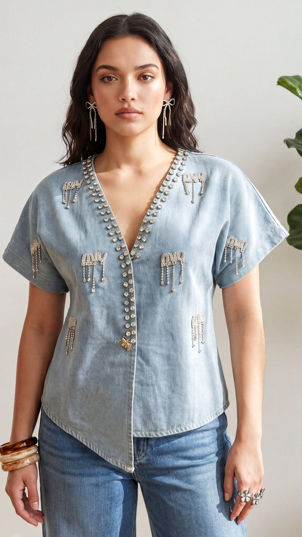 Woman wearing a light blue denim shirt with decorative elements and jeans, standing against a neutral background.