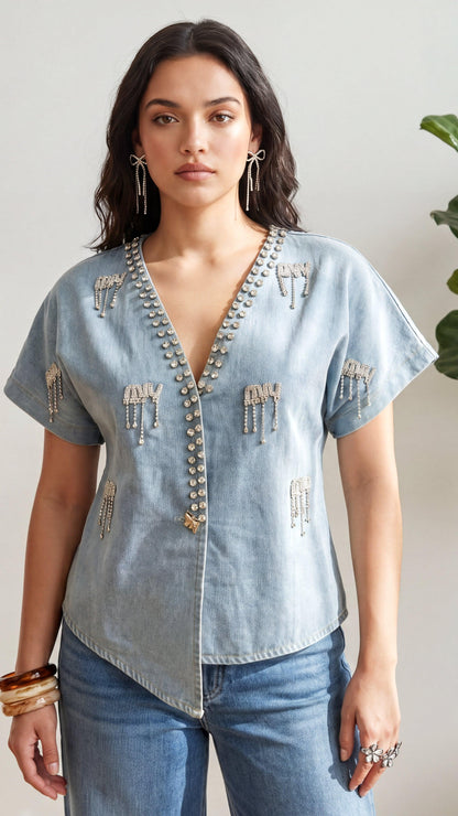 Woman wearing a light blue denim shirt with decorative elements and jeans, standing against a neutral background.
