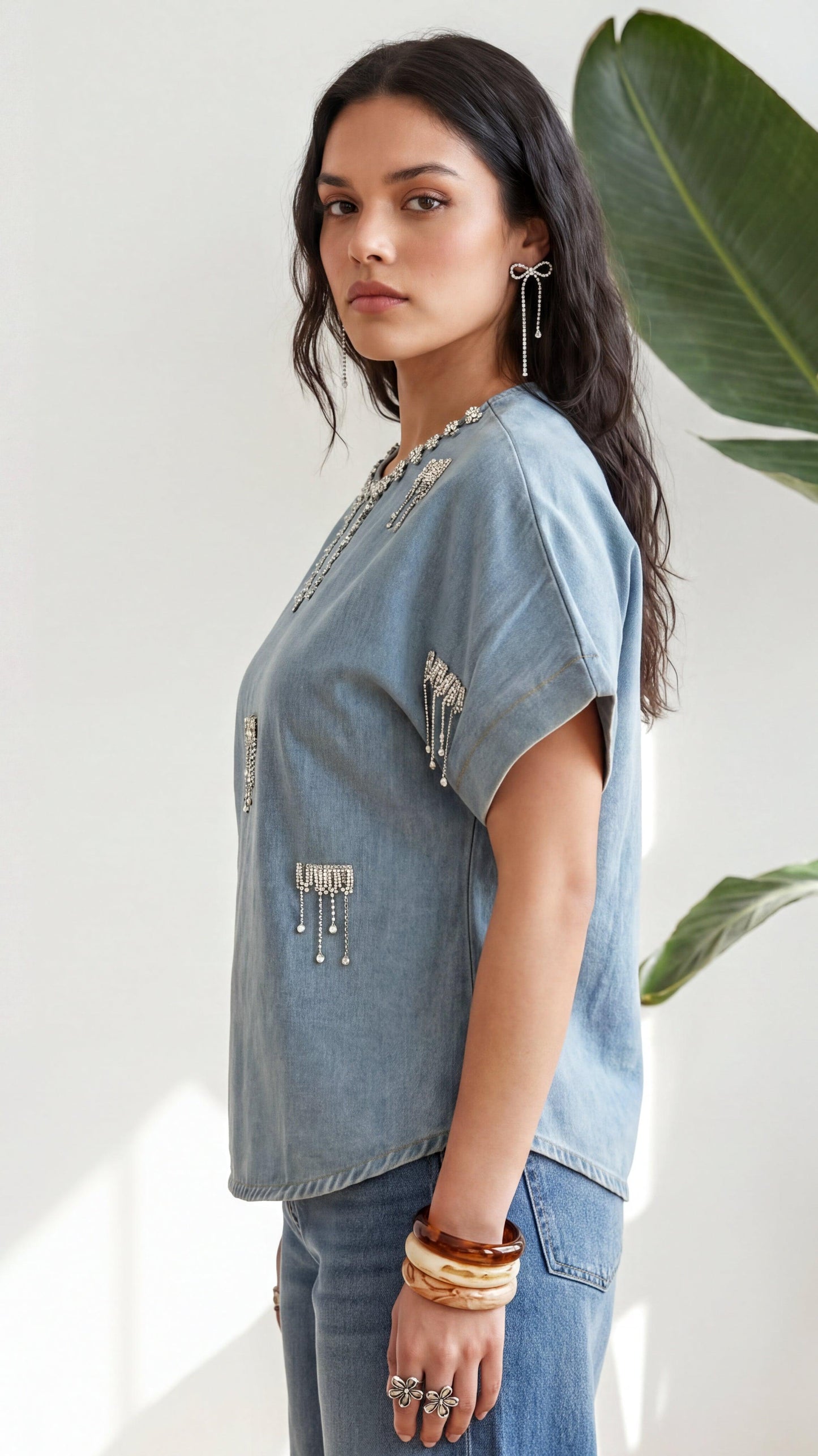 Woman wearing a denim top with decorative elements, standing indoors with a plant in the background.