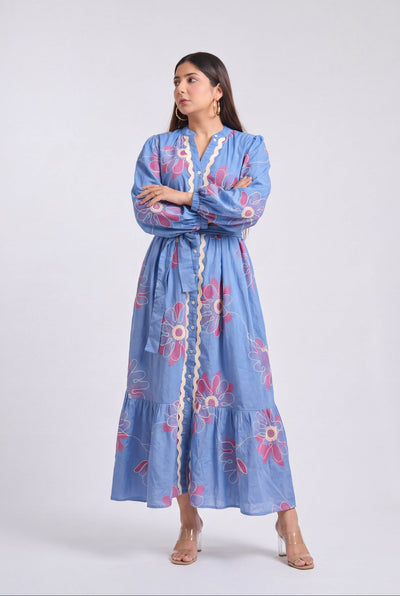 A woman modeling a light blue long-sleeve maxi dress with pink and white floral embroidery and a cream-colored button placket, standing against a light gray studio background.