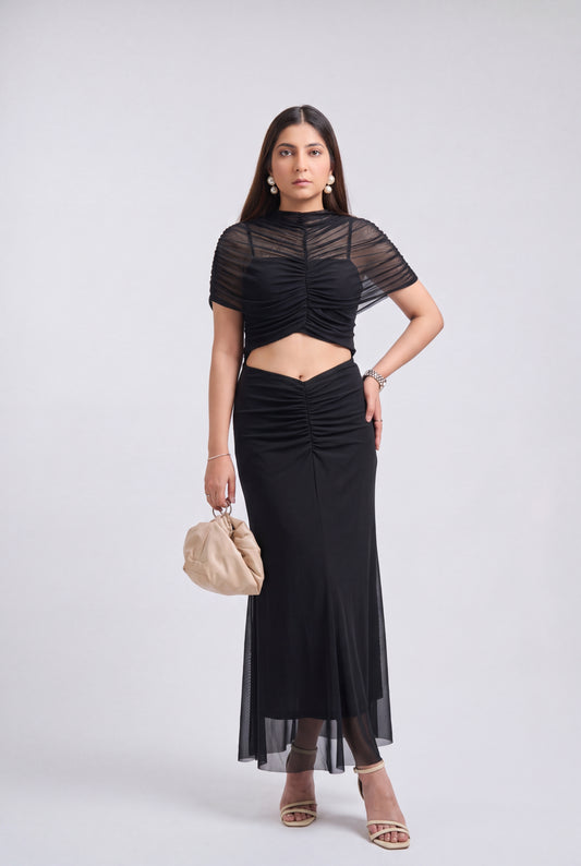 A woman modeling a black two-piece outfit consisting of a short-sleeve sheer mesh crop top and a matching high-waisted ruched midi skirt, paired with beige strappy heels and a beige handbag, against a light gray studio background.
