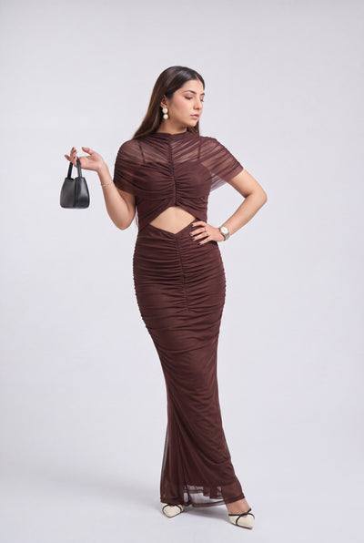 A woman modeling a brown ruched two-piece set consisting of a short-sleeve crop top with a cutout and a matching fitted maxi skirt, paired with black pointed-toe heels and a small black handbag, against a light gray studio background.