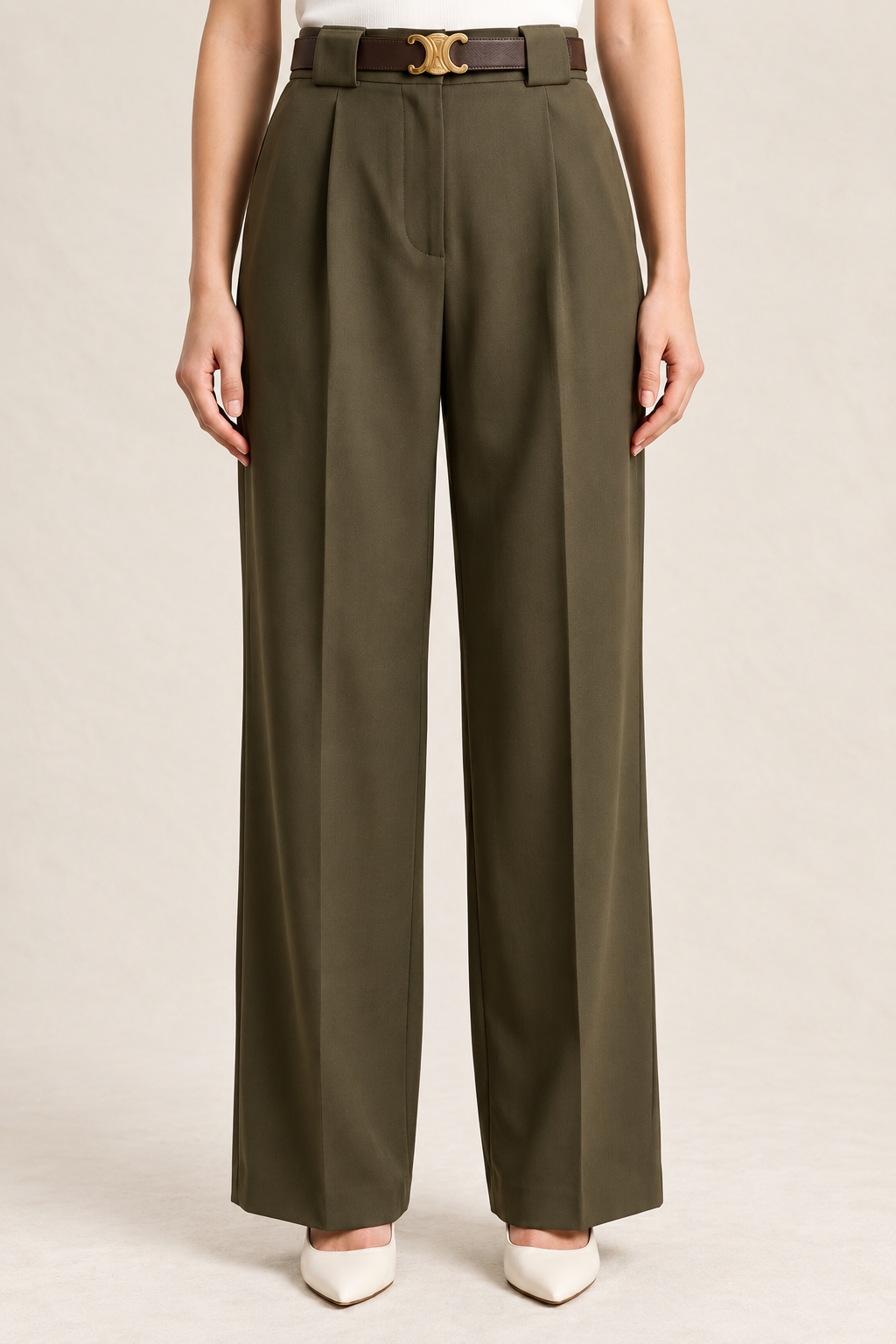 A model is wearing high-waisted, wide-leg olive green trousers with a dark brown leather belt featuring a gold-tone buckle, paired with white pointed-toe heels against a light beige studio background.