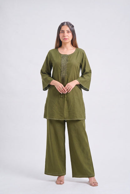 A woman modeling an olive green two-piece kurta set with wide-leg pants and three-quarter sleeves, standing against a light gray studio background.