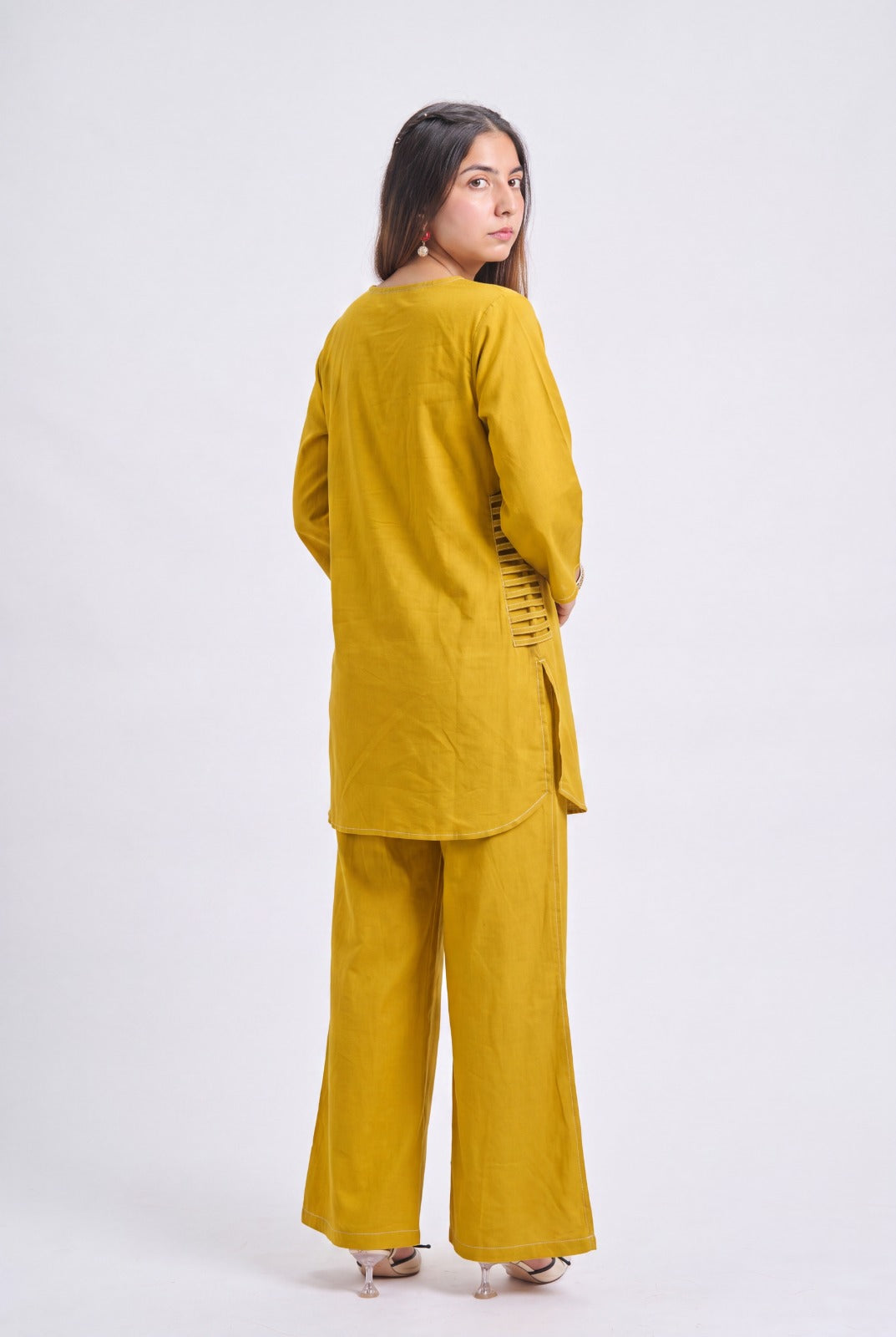 A woman modeling a mustard yellow two-piece linen set, featuring a long-sleeve tunic and wide-leg trousers, shown from a rear three-quarter angle against a light gray studio background.