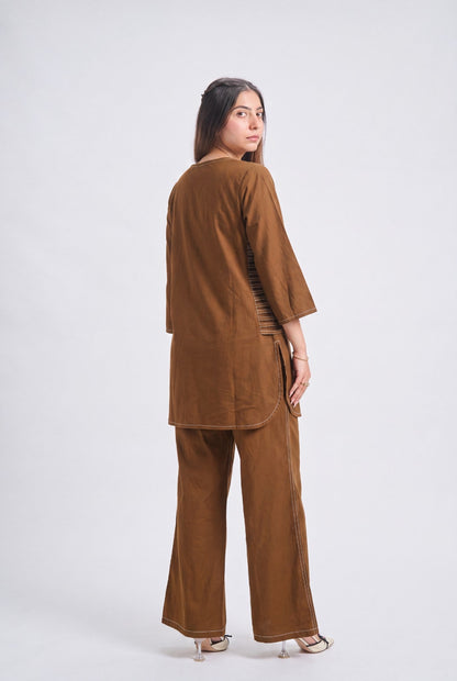 A woman modeling a brown two-piece linen set, featuring a short-sleeved tunic top and matching wide-leg trousers, shown from a rear three-quarter angle against a light gray studio background.
