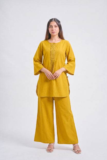 A woman modeling a mustard yellow two-piece kurta and trouser set against a plain light gray studio background.