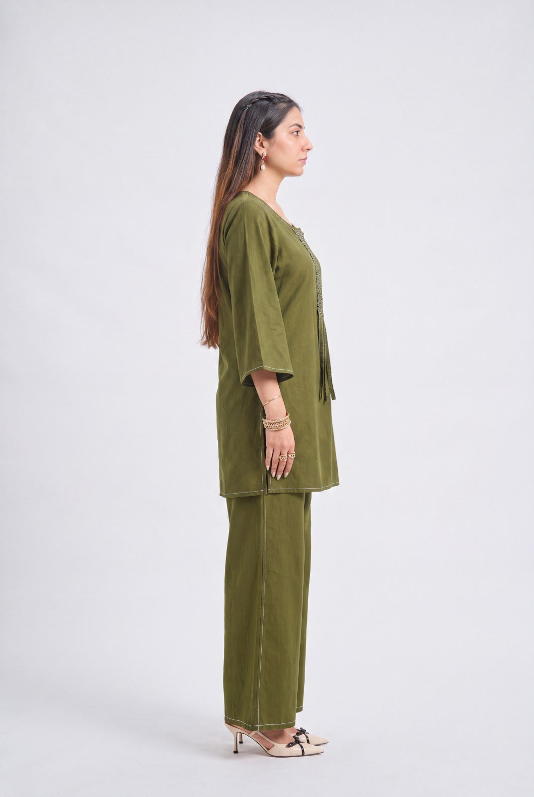 A woman stands in profile wearing an olive green two-piece outfit with a short tunic and wide-leg pants, styled with beige high-heeled sandals against a plain light gray studio background.