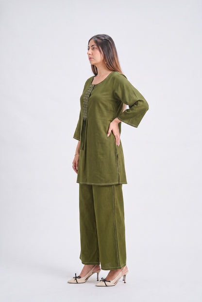 A woman modeling an olive green two-piece kurta and trouser set against a plain light gray studio background.